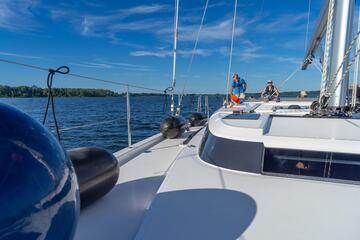 Im Vordergrund liegt ein Kugelfender, im Hintergrund stehen zwei Crewmitglieder am Ruder des Segelbootes.