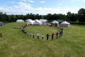 Junge Menschen stehen in einem Kreis und fassen sich an den Händen. Im Hintergrund stehen Zelte.