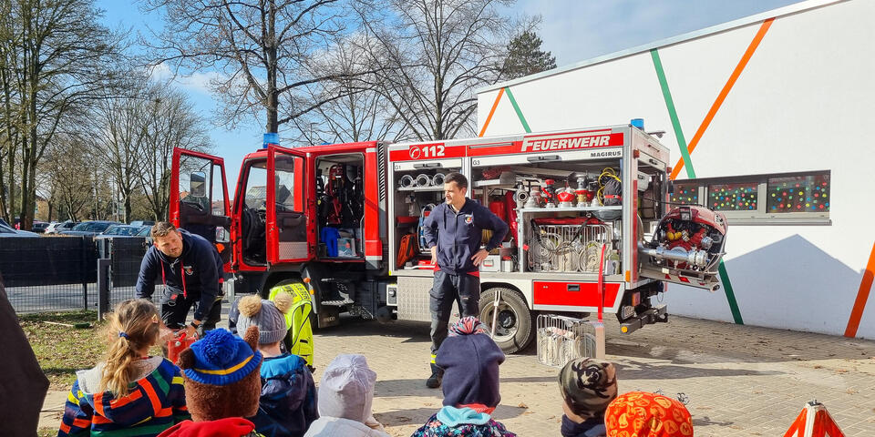 Eine Gruppe von Kindern schauen auf ein Feuerwehrauto. Davor steht ein Mann in Feuerwehrbekleidung