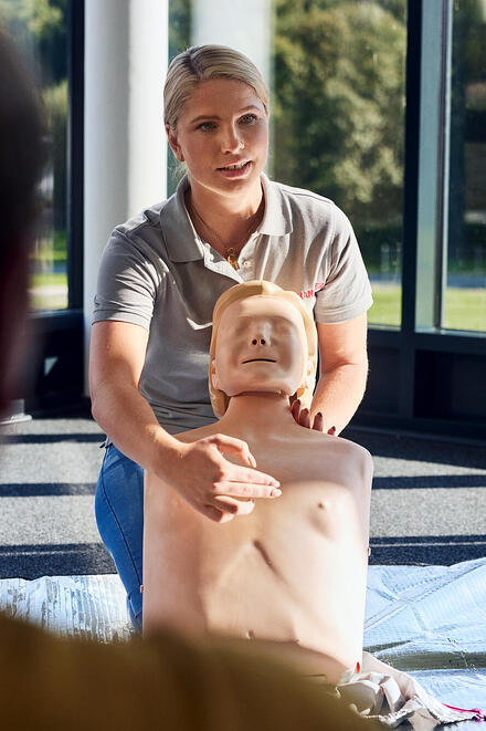 Eine Frau erklärt an einer Puppe die 1. Hilfe
