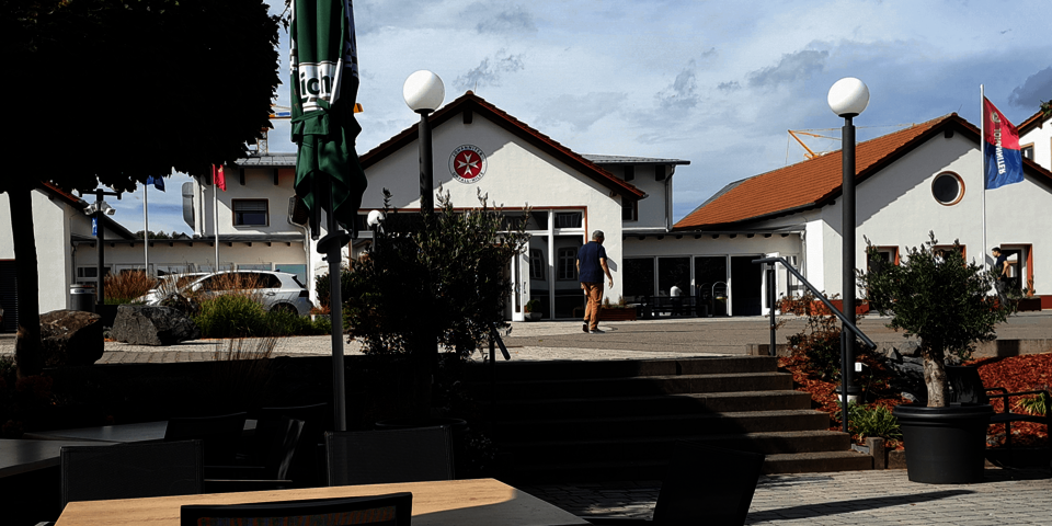 im Vordergrund eine Restaurantterrasse mit Tischen und Stühlen; im Hintergrund weißes Gebäude mit Johanniter-Zeichen am Giebel.