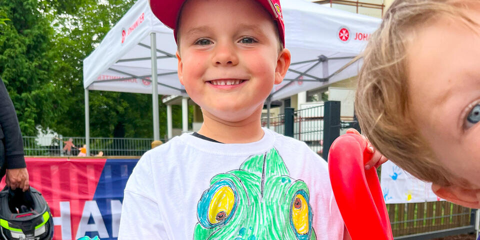 Ein kleiner Junge mit roter Baseballmütze und einem weißen T-Shirts auf dem ein grünes Chamäleon gemalt ist. Er hält einen roten Luftballon in Tierform