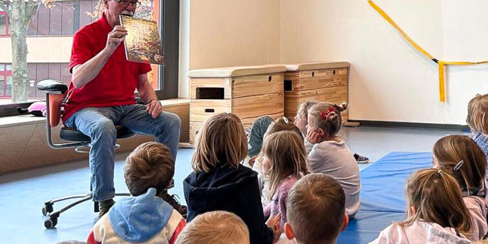 Ein Mann hält ein Buch und sitzt vor einer Gruppe von Kindern und Erwachsenen