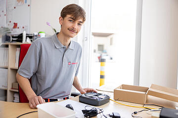 Ein junger Mann in einem grauen Poloshirt mit dem Logo der Johanniter. Er sitzt an einem Schreibtisch und arbeitet mit einem technischen Gerät, umgeben von Kabeln und kleinen Boxen. Im Hintergrund ist ein heller Raum mit einem Fenster zu sehen, durch das Tageslicht einfällt. Die Stimmung wirkt freundlich und konzentriert.