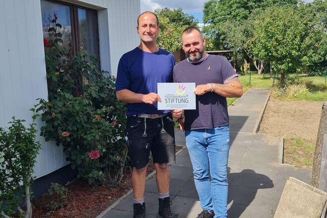 Man sieht zwei Personen in einem Garten stehen. Dahinter ein Gartenhaus.