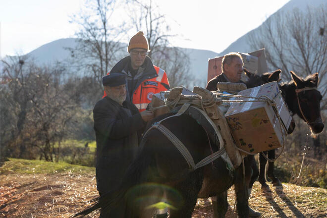 Drei Männer beladen in einer ländlichen Berglandschaft einen Esel mit Kisten, während einer in einer orangefarbenen Einsatzjacke daneben steht.