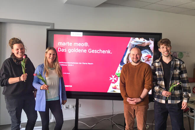 Zwei Frauen mit einer Tulpe in der Hand und zwei Männer, einer hält ebenfalls eine Tulpe in der Hand. Hinter ihnen ein Bildschirm auf dem zu lesen ist "Marte Meo - ein Geschenk"