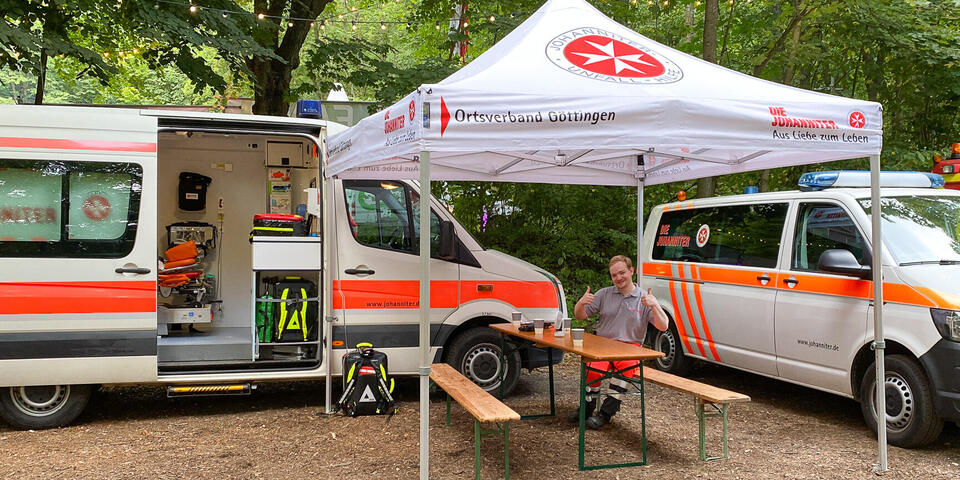 Es sind Johanniter-Einsatzfahrzeuge zu sehen. Unter einem Pavillons sitzt ein Johanniter.