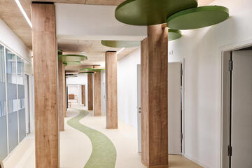 Ein Flur. Am Boden schlängelt sich eine geschwungene grüne Bahn. Holzpfeiler sind zu sehen. An der Decke bei den Holzpfeilern sind grüne Kreise.