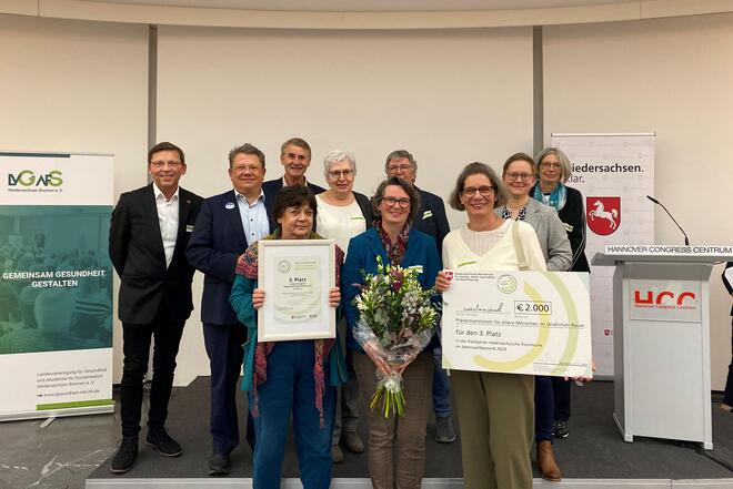 Menschen auf einer Bühne. Im Vordergrund hält eine Frau eine Urkunde hoch, daneben eine Frau mit einem Blumenstrauß und rechts eine Frau mit einem symbolischen Scheck.