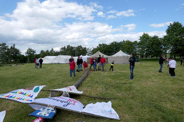 Ein Mast liegt auf einer Wiese. An diesem Mast sind verschiedene Banner befestigt. Im Hintergrund stehen Zelte.