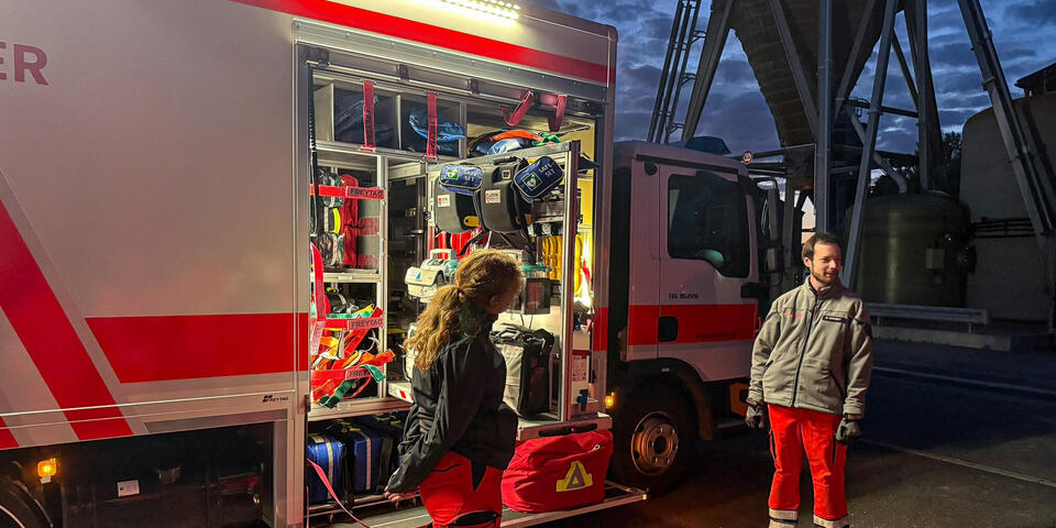Ein Mann und eine Frau in Einsatzkleidung vor einem großen Gerätewagen. Die Seitentür des Fahrzeugs ist offen und man sieht viele Gerätschaften