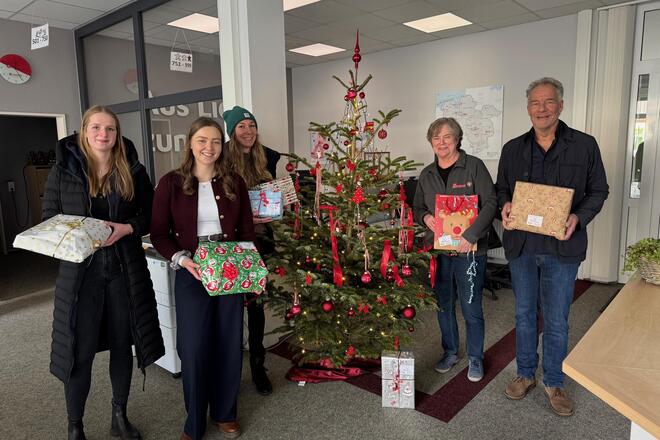 Fünf Menschen mit festlich verpackten Geschenken stehen vor einem Weihnachtsbaum.