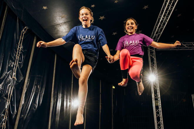 Zwei fröhliche Kinder springen in die Luft, halten sich an den Händen und tragen T-Shirts mit dem Schriftzug "LILALU". Sie befinden sich in einem Zirkuszelt mit Sternen an der Decke, und Lichter sorgen für eine dynamische Atmosphäre.
