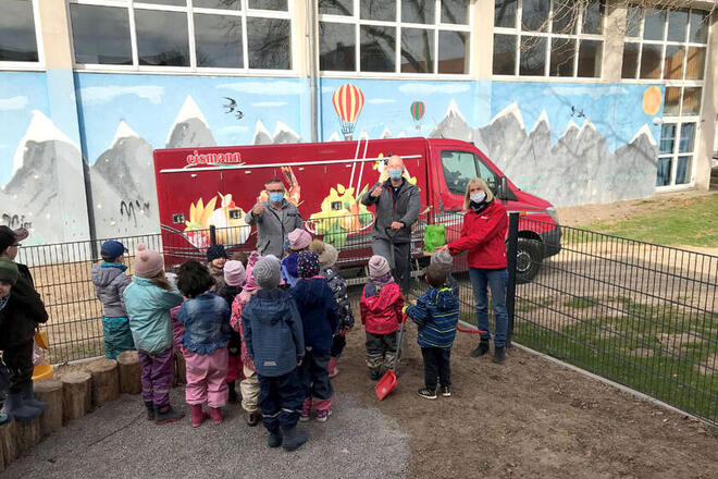 Kita-Kinder, Dienstellenleiter und Kita-Leitung pflanzen gemeinsam mit einem eismann-Mitarbeiter einen kleinen Baum, im Hintergrund steht der Lieferwagen der Firma eismann. 