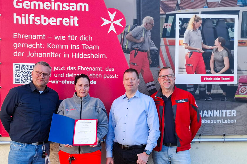 Eine Frau in Johanniter-Kleidung hält eine Urkunde hoch. neben ihr drei Männer. Alle stehen an einer Plakatwand auf der Johanniter und die Worte "Gemeinsam hilfsbereit" zu sehen sind.