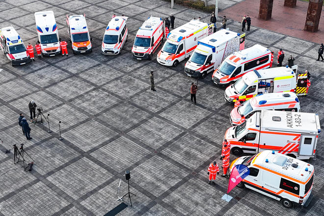 Man sieht mehrere Krankentransportwagen der Johanniter in Rot-Orange, die im Halbkreis stehen, und Menschen in Einsatzkleidung, die neben den Wagen stehen. Dazwischen kann man vereinzelt Menschen in dunkler Kleidung sehen. 