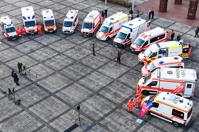 Man sieht mehrere Krankentransportwagen der Johanniter in Rot-Orange, die im Halbkreis stehen, und Menschen in Einsatzkleidung, die neben den Wagen stehen. Dazwischen kann man vereinzelt Menschen in dunkler Kleidung sehen. 