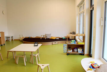 Blick in den Gruppenraum. Man sieht ein kleinen Tisch mit Minihockern, einen Bau- und Spielteppich sowie einige Spielsachen. 