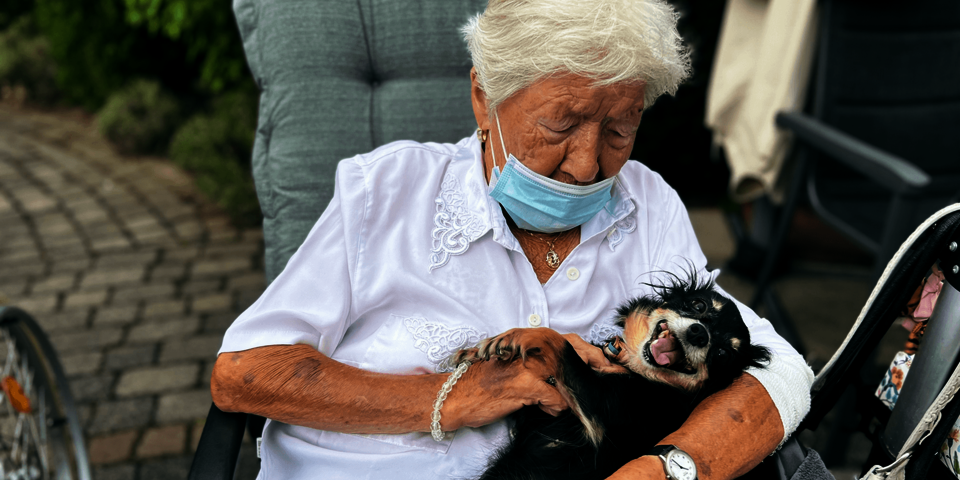 Eine weißhaarige Frau sitzt in einem Gartenstuhl. Sie trägt eine weiße Bluse und streichelt einen kleinen Hund.