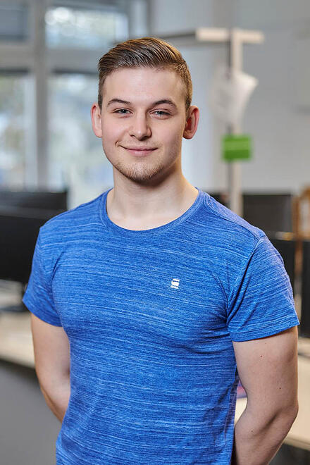 Ein junger Mann in blauem T-Short steht in einem Büro und blickt in die Kamera