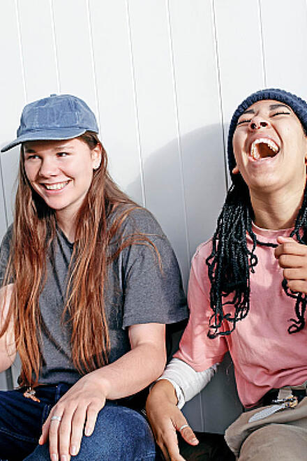 Zwei FSJlerinnen sitzen lachend nebeneinander vor einer weißen Wand