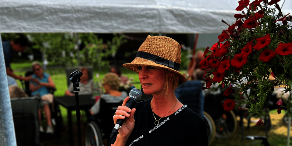 Ein junge Frau mit Strohhut hält in einem Garten ein Mikrofon; im Hintergrund Gartentische