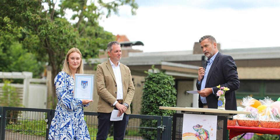 Ein Mann mit Mikrophon an einem Rednerpult ein Mann und eine Frau, die eine Urkunde hochhält hören ihm zu. 