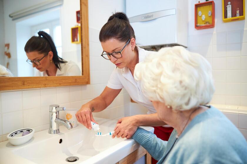 Eine Mitarbeiterin der Johanniter hilft einer Seniorin dabei, Zahnpasta auf die Zahnbürste zu geben.