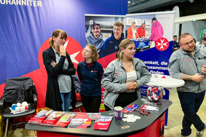 Drei Frauen und ein Mann in Johanniter-Kleidung an einem Messestand der Johanniter in einer Halle