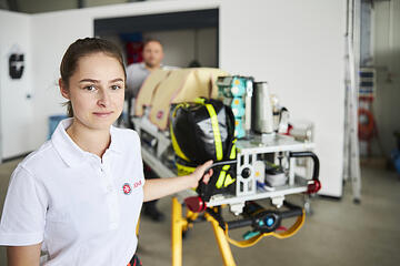 Eine junge Frau in einem weißen Poloshirt mit dem Johanniter-Logo. Sie steht neben einer Fahrtrage mit medizinischem Equipment. Im Hintergrund ist ein Mann zu erkennen, der unscharf dargestellt ist.