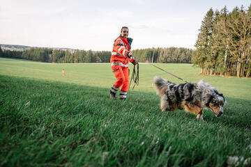 Eine Frau in auffälliger Einsatzkleidung hält einen Rettungshund an der Leine. Der Hund schnüffelt konzentriert im Gras, während ein zweiter Einsatzhelfer weiter entfernt auf der Wiese steht. Im Hintergrund ist ein Wald zu sehen.