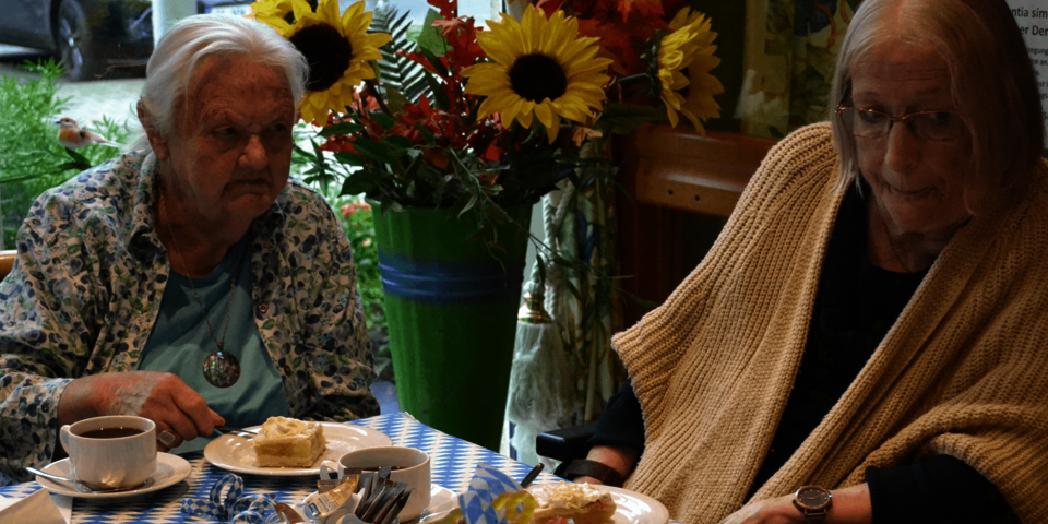 zwei ältere Damen sitzen am blauweiß gedeckten Kaffeetisch, im Hintergrund Sonnenblumen