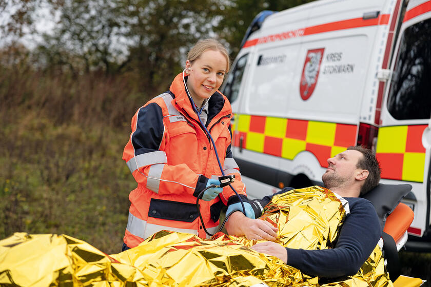 Eine Ehrenamtliche des Katastrophenschutzes betreut einen Patienten, der mit einer Rettungsdecke zugedeckt ist. Im Hintergrund steht ein Fahrzeug des Katastrophenschutzes.