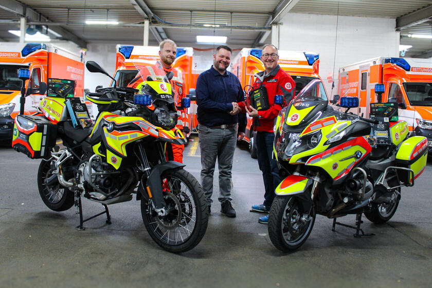 Zwei Johanniter mit ihren Motorrädern in einer Fahrzeughalle. Zwischen ihnen steht ein Mann, der einem Johanniter die Hand schüttelt. Im Hintergrund sind Rettungswagen.