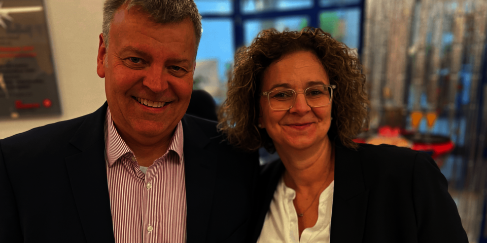 Ein Mann und eine Frau, beide in dunklen Blazern, stehen nebeneinander und lächeln in die Kamera
