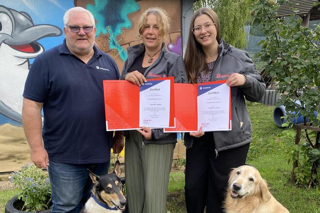 Drei Erwachsene stehen mit zwei Hunden vor einer bunt bemalten Wand im Garten der Kita Hoppetosse in Schwabach. Zwei der Erwachsenen halten Zertifikate in den Händen. Die beiden Hunde, ein Collie-Mischling und ein Golden Retriever, sitzen entspannt auf dem Rasen.