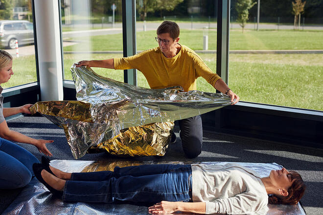 Erste-Hilfe-Kurs in hellem Raum, Frau wird mit Goldfolie vor Kälte geschützt.