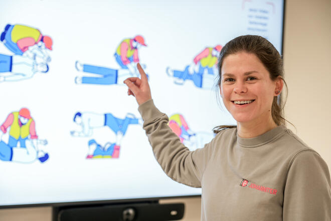 Lächelnde Ausbilderin deutet auf Bildschirm mit Anleitungen zur stabilen Seitenlage.