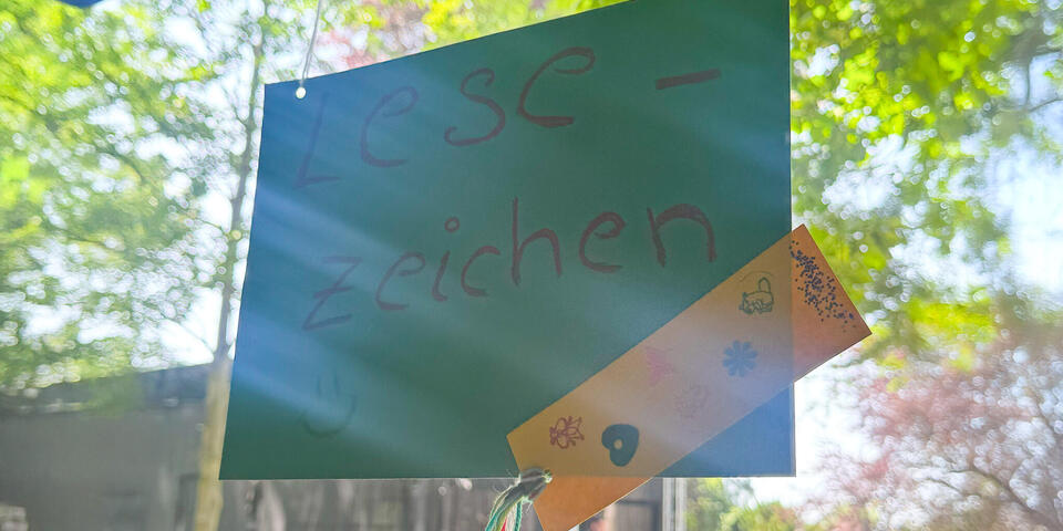 Ein blaues Pappschild auf dem steht "Lesezeichen". Im Hintergrund ein Park mit Menschen