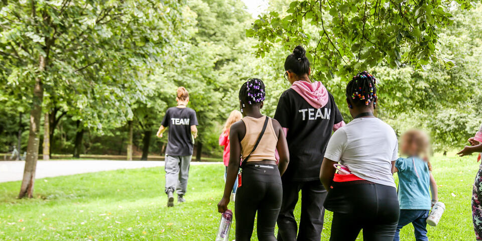 Rückansicht einer Gruppe bei der Ferienbetreuung; Betreuende führen Kinder über die Wiese.