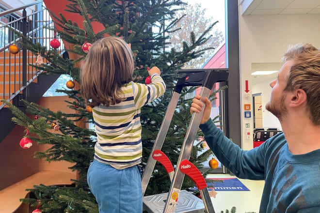 Ein kleines Mädchen steht auf einer Leiter und befestigt Weihnachtsschmuck an einer großen Tanne. Ein Mann hält sie fest.