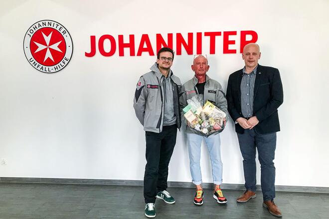 Drei Männer vor einer Wand auf dem das Johanniter-Logo steht. Der Mann in der Mitte hält einen Präsentkorb