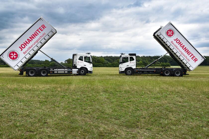Zwei große weiße Logistik-LKW stehen sich mit hochgeklapptem Container gegenüber. Auf der Plane des Containers ist das Logo der Johanniter zu sehen.