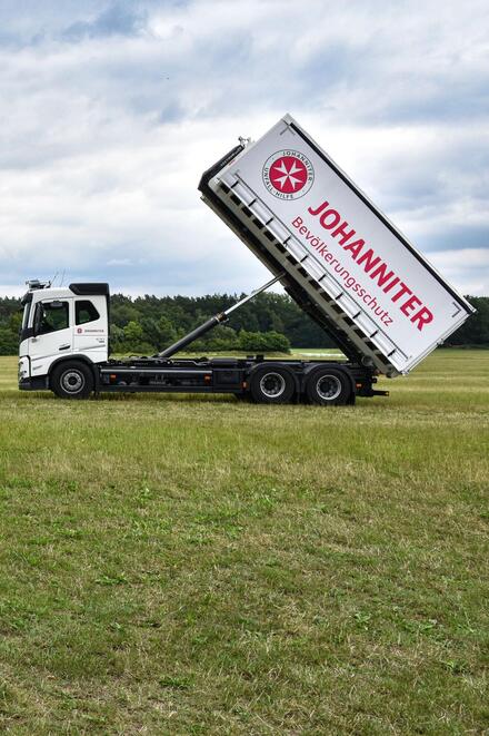 Zwei große weiße Logistik-LKW stehen sich mit hochgeklapptem Container gegenüber. Auf der Plane des Containers ist das Logo der Johanniter zu sehen.