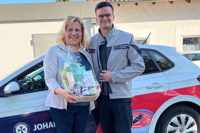 Eine Frau mit einem Geschenkkorb und ein Mann in Johanniter-Kleidung vor einem Johanniter-Auto