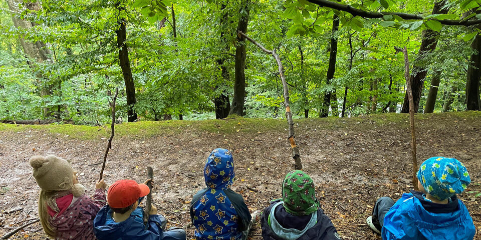Fünf Kinder in einem Wald. Sie haben Äste in den Händen und spielen Angeln.