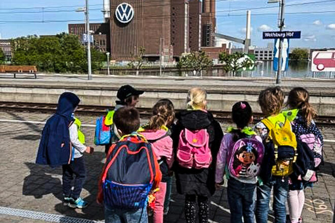 Eine Gruppe von Kindern auf einem Bahnsteig. Im Hintergrund ein VW-Werk.