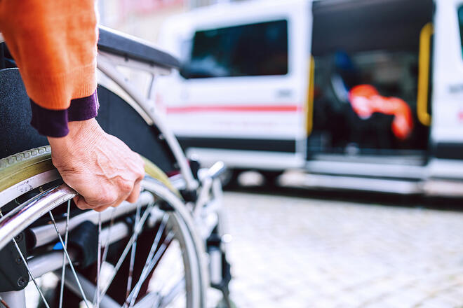 Eine Hand greift an das Rad eines Rollstuhls, im Hintergrund ist ein geöffneter Johanniter-Fahrdienstwagen zu sehen.