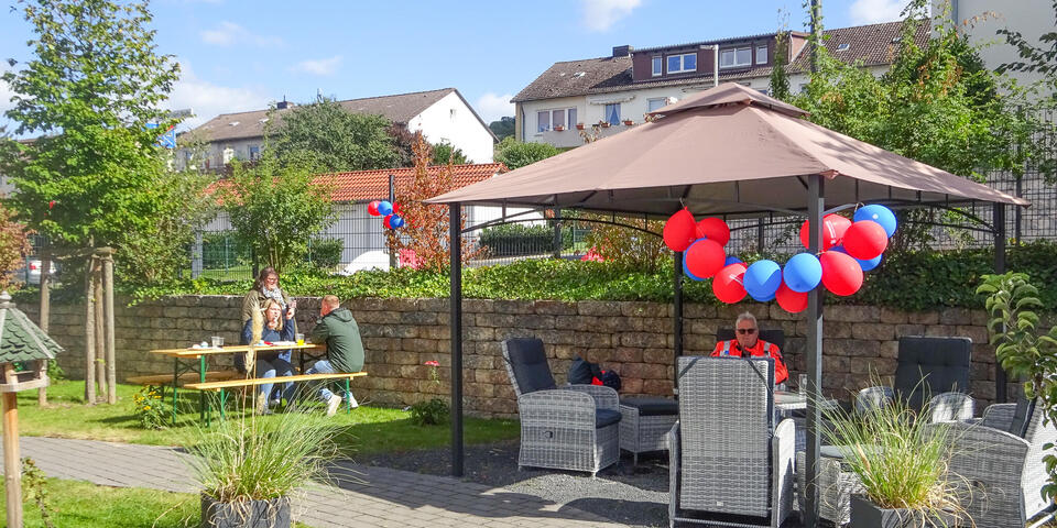 Ein Pavillon im Garten, der mit Luftballons geschmückt ist. Unter dem Pavillon sitzen Menschen.